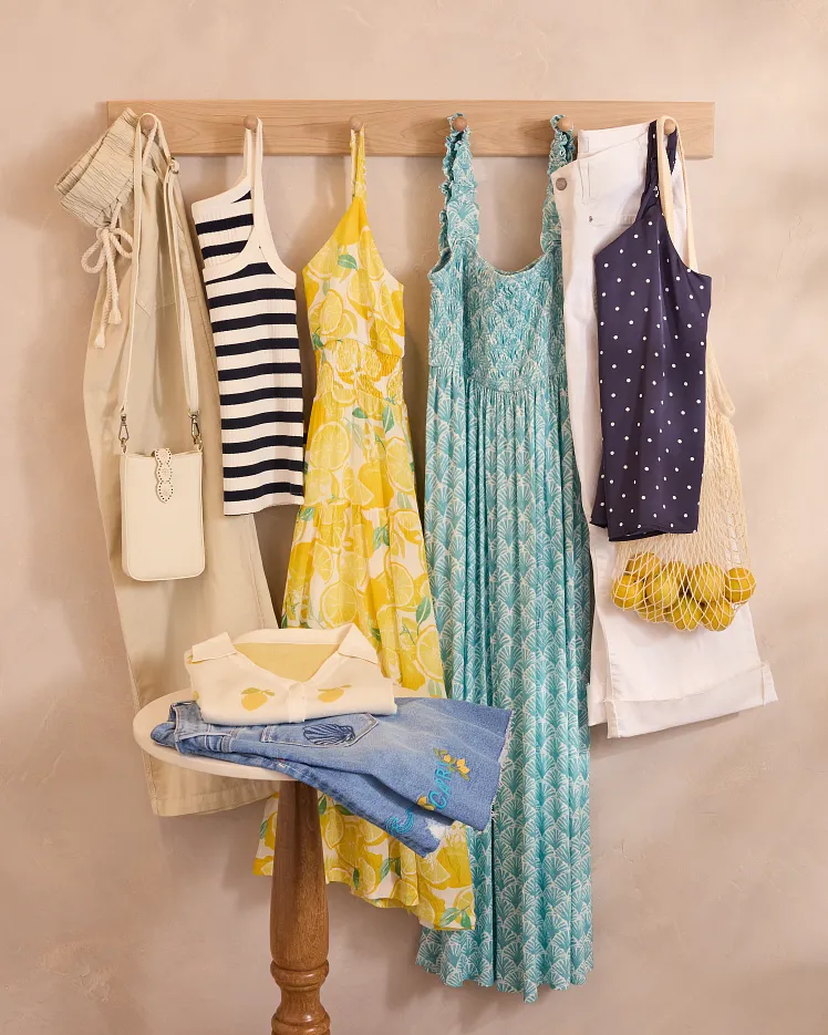 Green floral sleeveless dress draped over a modern chair and styled with a brown belt, light blue blazer, and sunglasses on the seat. The outfit is arranged as a complete warm weather look against a neutral background to showcase the styling pieces together.