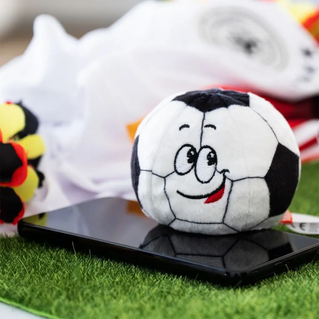 Kleiner, weicher Pl&uuml;sch-Fu&szlig;ball Schmoozies&reg; Classic mit lachendem Gesicht liegt auf einem Smartphone-Display auf Kunstrasen &ndash; Emotionaler WM-Werbeartikel.