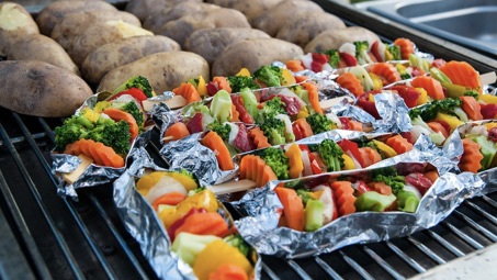 Warzywne szaszłyki ułożone na folii aluminiowej oraz ziemniaki pieczone bezpośrednio na grillowym ruszcie