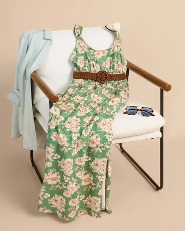Green floral sleeveless dress draped over a modern chair and styled with a brown belt, light blue blazer, and sunglasses on the seat. The outfit is arranged as a complete warm weather look against a neutral background to showcase the styling pieces together.