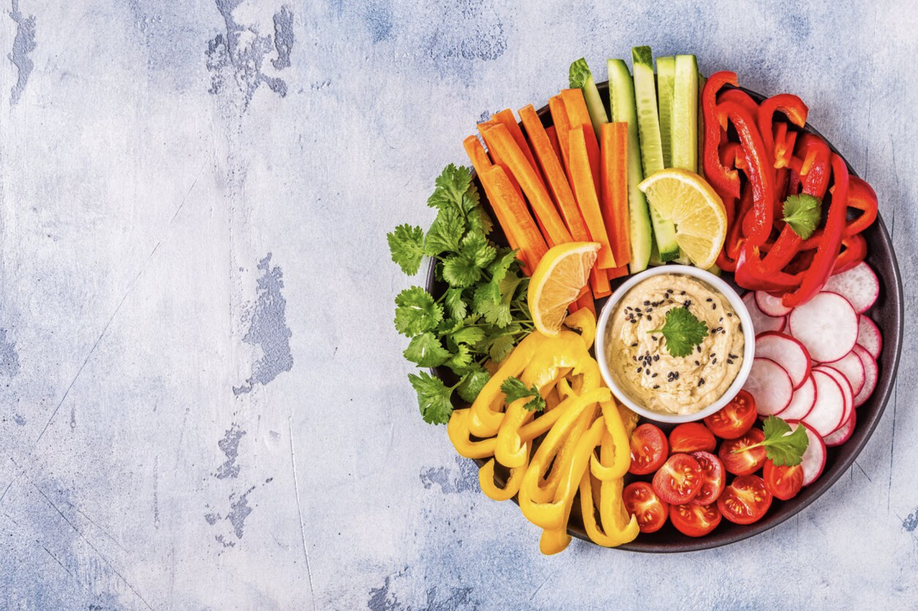 okrągła miseczka hummusu podana na talerzu wypełnionym świeżymi warzywami.