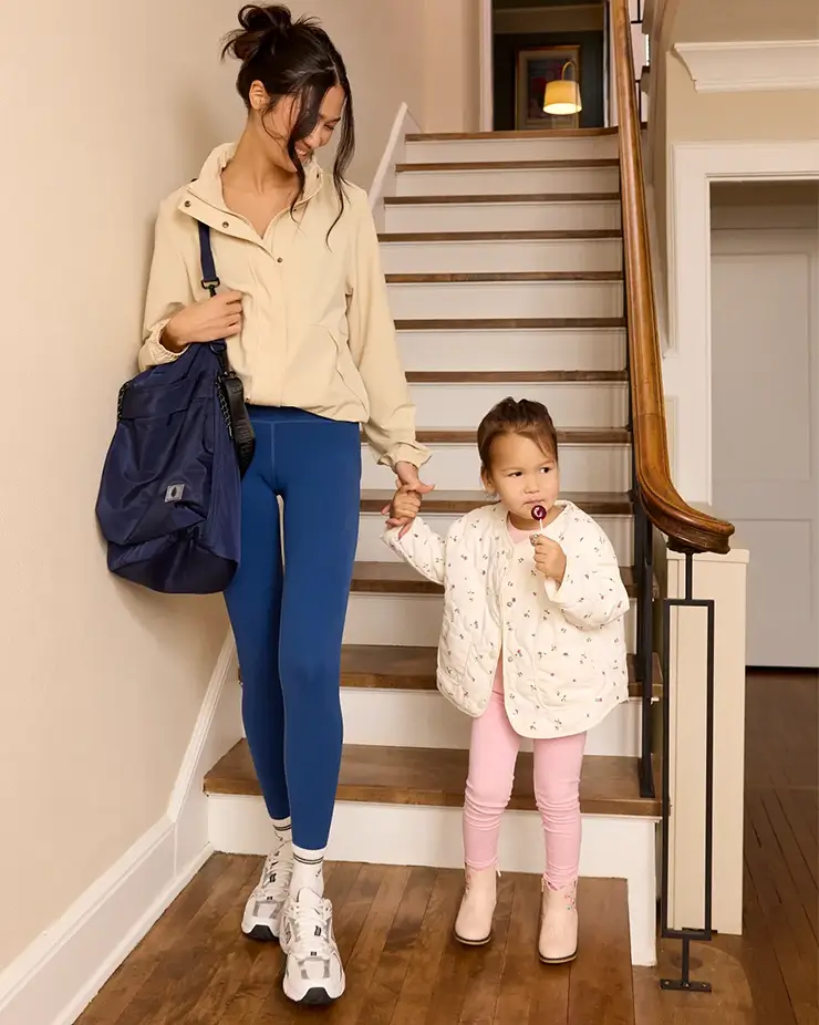 Hardshell suitcase with brown trim topped with a stack of women&rsquo;s clothing and a blue hat that reads "Salty". A larger folded stack of outfits sits in the lower left corner beside gold jewelry including a charm necklace and earrings styled as part of the travel wardrobe display.