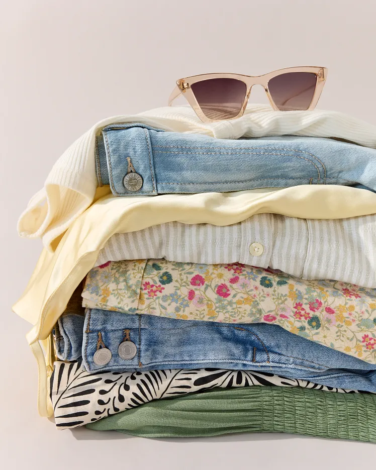 Green floral sleeveless dress draped over a modern chair and styled with a brown belt, light blue blazer, and sunglasses on the seat. The outfit is arranged as a complete warm weather look against a neutral background to showcase the styling pieces together.