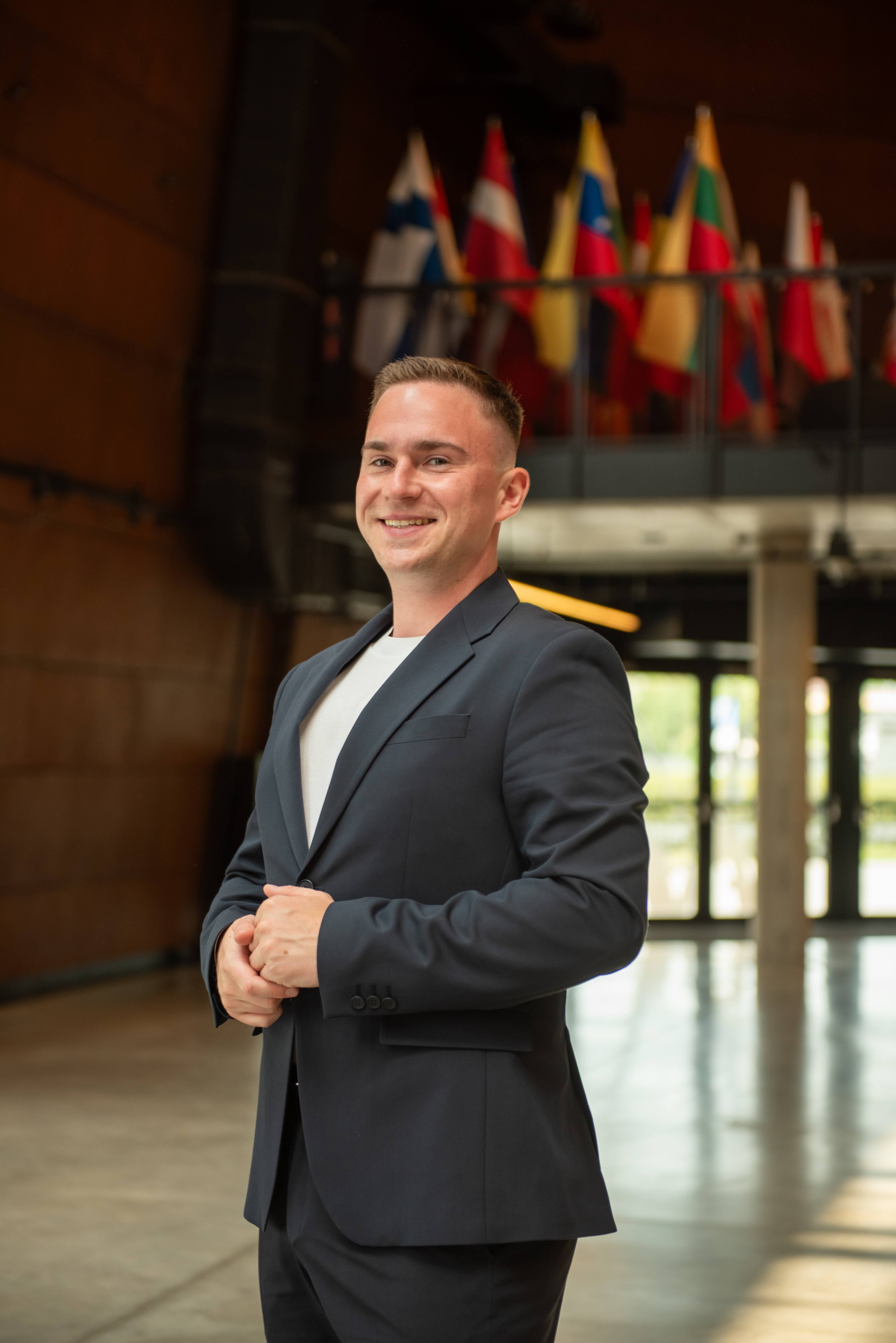 Oskar Król – konsultant marketingowo-sprzedażowy, specjalista od rynków zagranicznych Oskar Król w garniturze na tle flag międzynarodowych – ekspert ds. ekspansji zagranicznej