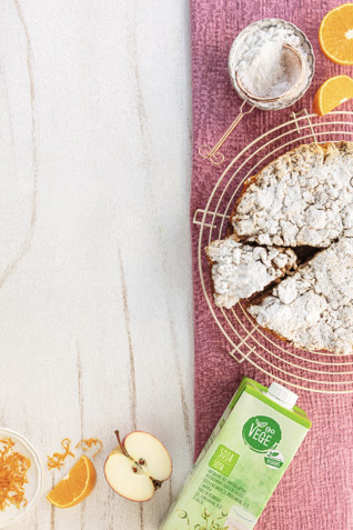 Zielony karton z napojem sojowym GoVege, ułożony obok świeżych owoc&oacute;w pomarańczy i jabłka oraz kruchego ciasta posypanego cukrem pudrem