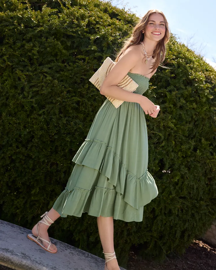 Green floral sleeveless dress draped over a modern chair and styled with a brown belt, light blue blazer, and sunglasses on the seat. The outfit is arranged as a complete warm weather look against a neutral background to showcase the styling pieces together.