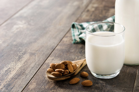 Szklanka napoju roślinnego, obok kt&oacute;rej leżą migdały na drewnianej łyżce, w tle ściereczka w zielono-białą kratę.