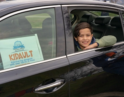 Boy in a car smiling with the Kidault logo sign.