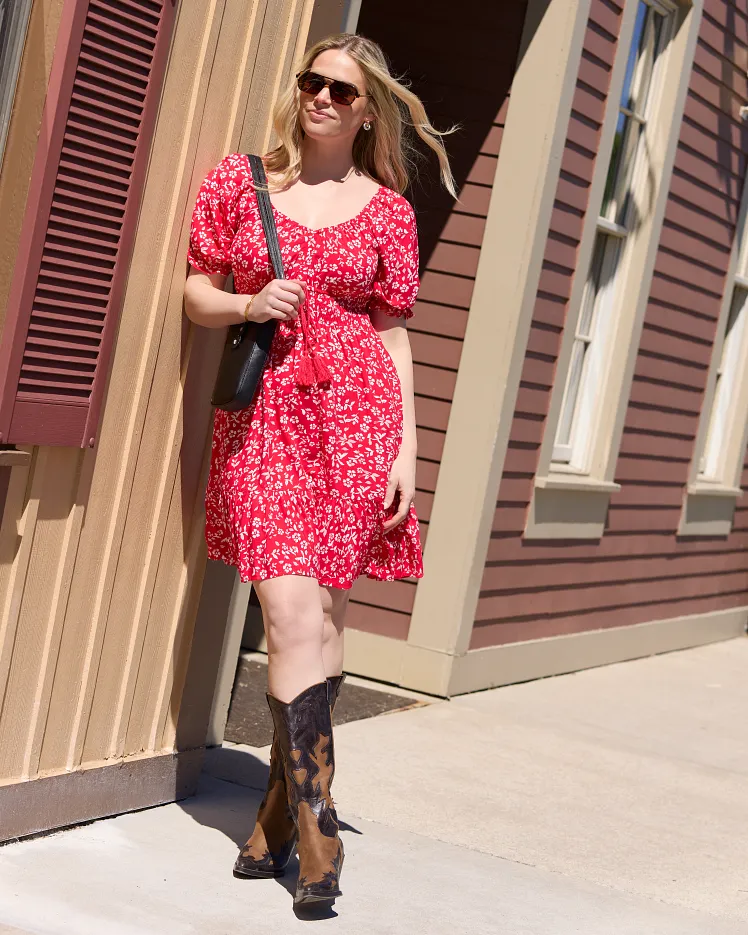 Green floral sleeveless dress draped over a modern chair and styled with a brown belt, light blue blazer, and sunglasses on the seat. The outfit is arranged as a complete warm weather look against a neutral background to showcase the styling pieces together.