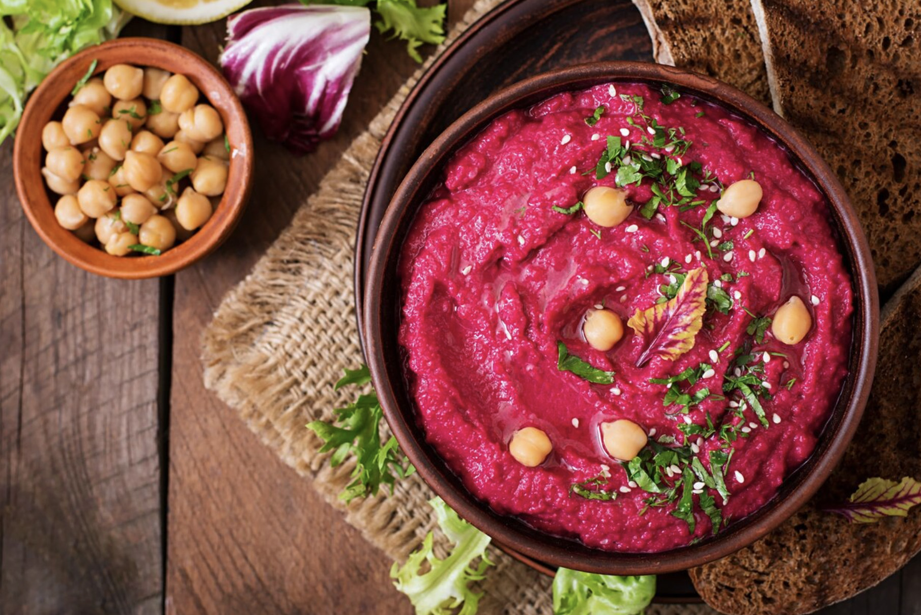 Hummus z burakiem, obsypany świeżymi ziołami i ziarnami ciecierzycy, podany w brązowej misce. Obok mniejsza miseczka, wypełniona nasionami cieciorki.